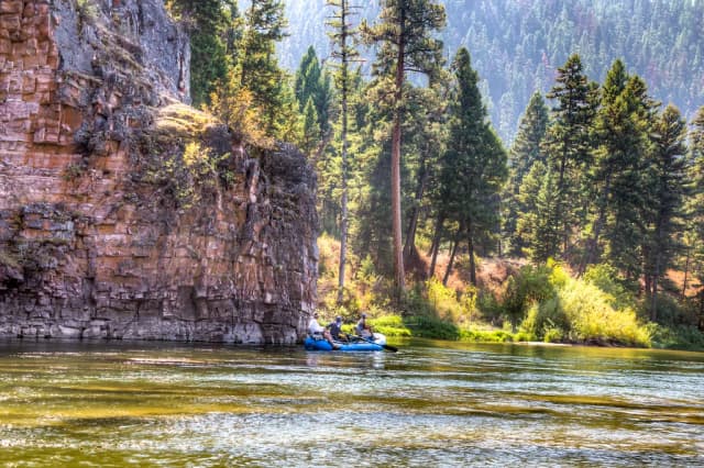 A raft with three anglers floats the Blackfoot River past a rock cliff and pine forest near Missoula, Montana.