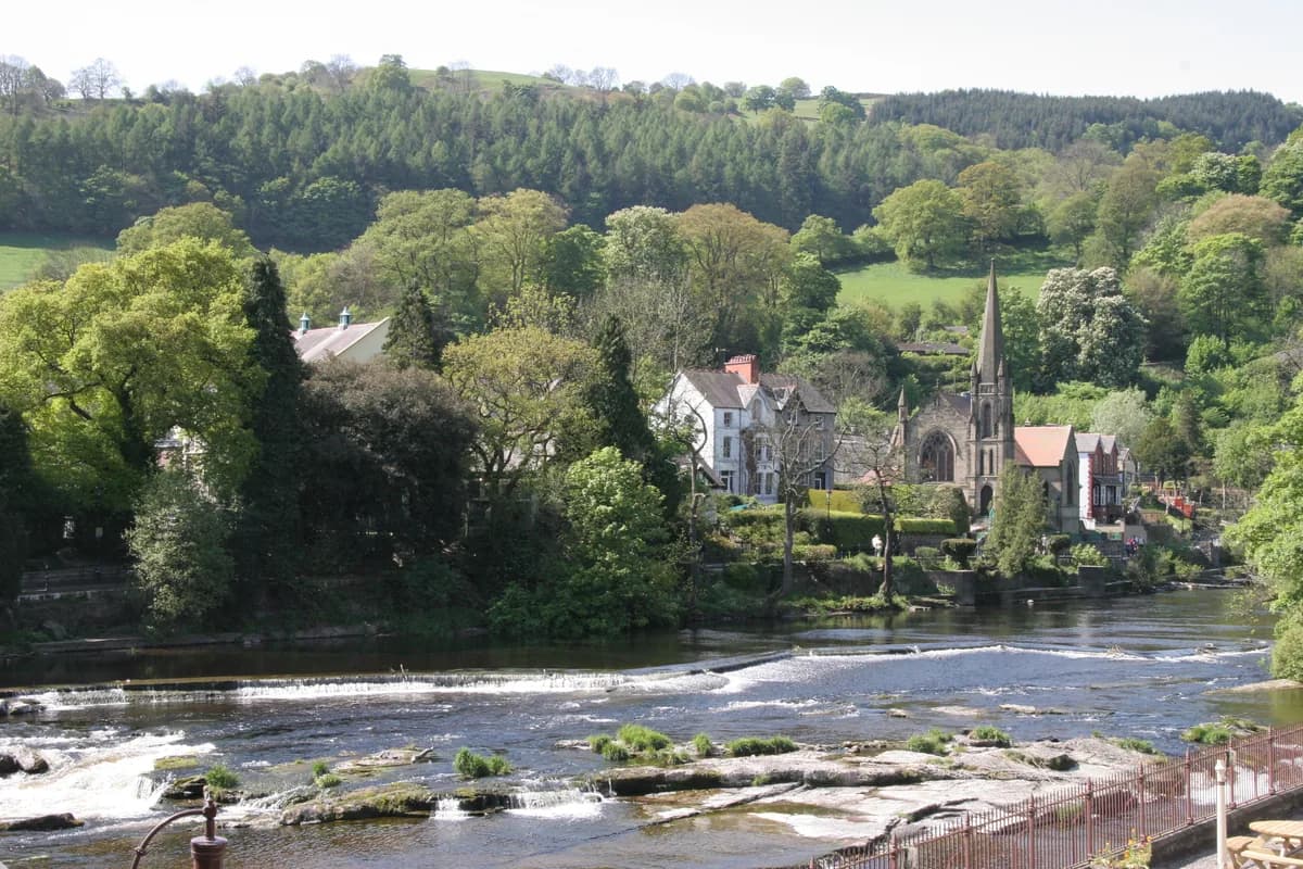 Wide view of the River Dee flowing over a weir through the town of Llangollen, Denbighshire, Wales, with wooded hills behind