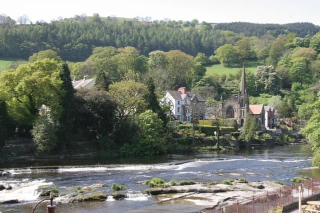 Wide view of the River Dee flowing over a weir through the town of Llangollen, Denbighshire, Wales, with wooded hills behind