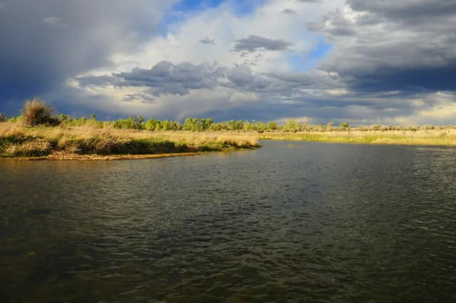 A wide, calm stretch of the Green River bordered by grassy banks under a dramatic stormy sky in Wyoming.