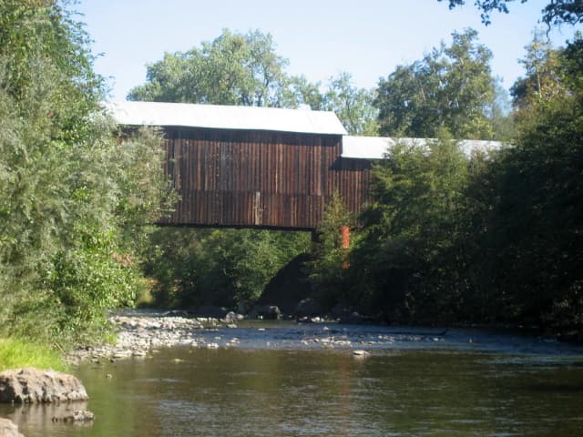 A wooden covered bridge crossing Butte Creek, with a gravel bar and calm water in the foreground, near Chico, California.