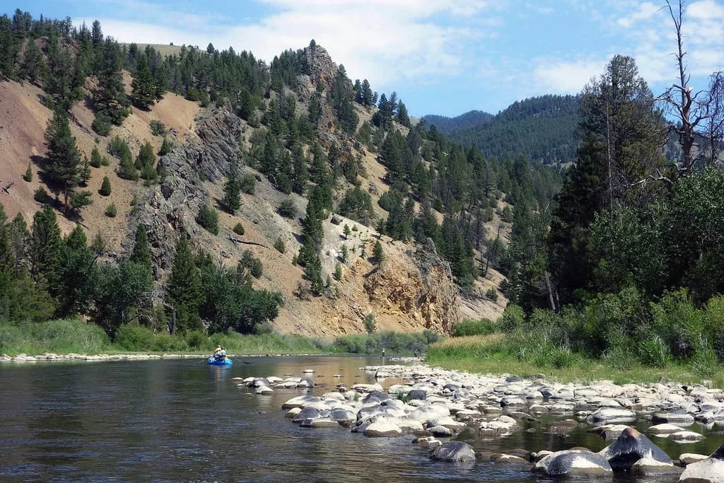 A drift boat floats past wading fly anglers on the Big Hole River, with a steep pine-and-rock canyon wall behind, Montana.