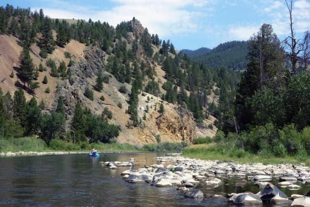 A drift boat floats past wading fly anglers on the Big Hole River, with a steep pine-and-rock canyon wall behind, Montana.