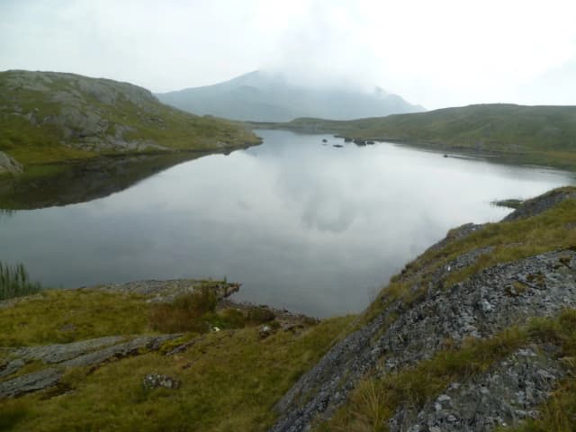 Calm upland lake (Llyn Cwm Corsiog) surrounded by rocky, grassy banks with misty hills in the background, Snowdonia.