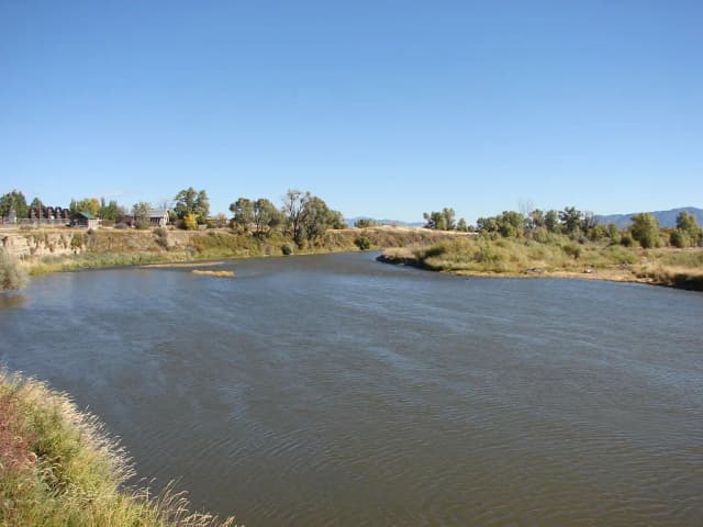 A broad, calm stretch of the Jefferson River bordered by grassy banks and cottonwood trees, with mountains in the distance.