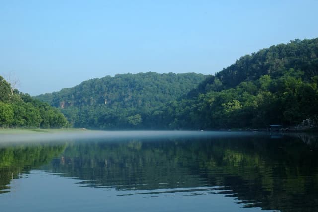 Calm White River in Arkansas at dawn, with mist rising off the water and wooded Ozark hills along the far bank.
