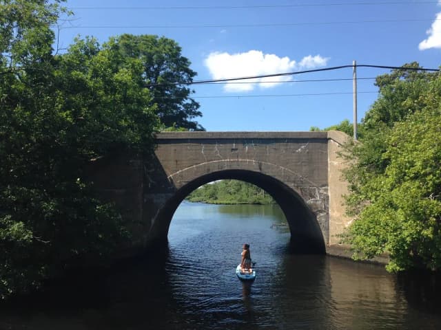 The Carmans River flowing under an old stone arch bridge on Long Island, New York, with a paddleboarder on the water and trees lining both banks.