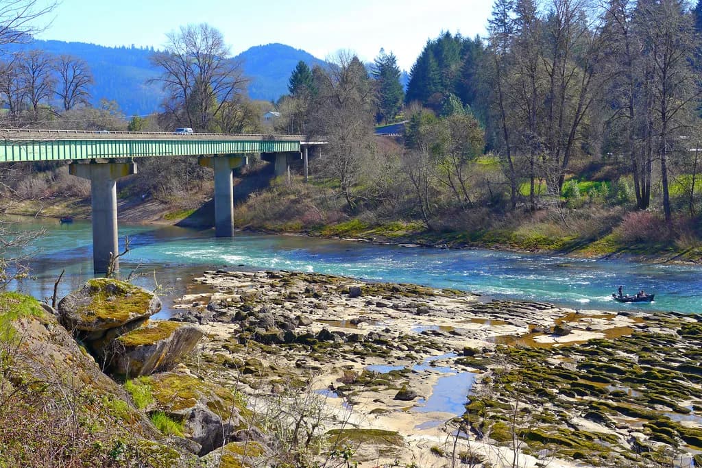 A fishing boat on the turquoise North Umpqua River beneath a highway bridge, with forested hills in the background.