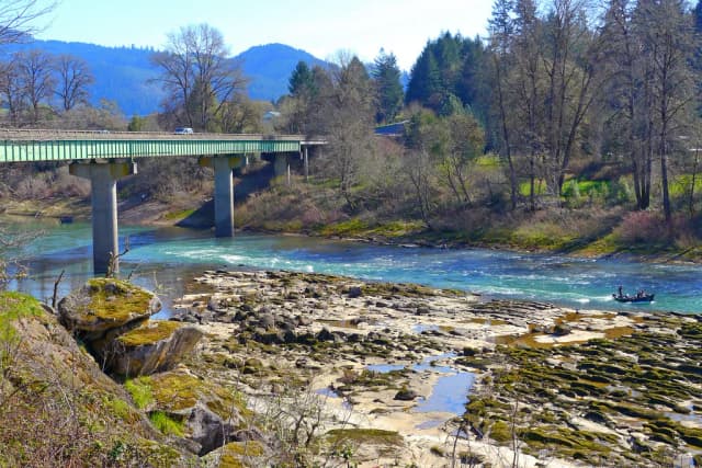 A fishing boat on the turquoise North Umpqua River beneath a highway bridge, with forested hills in the background.