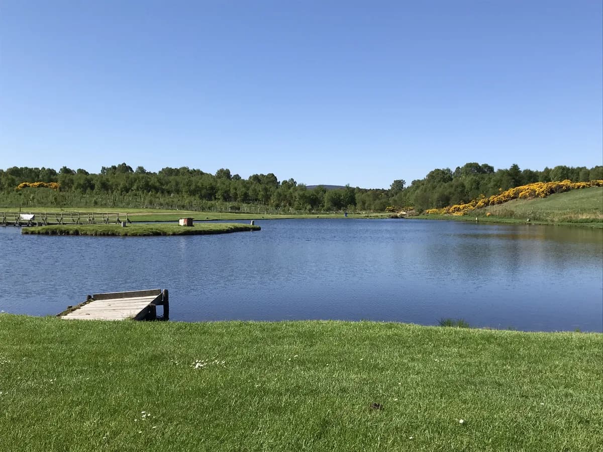 A stocked fishing lake at Achagour with a wooden angling platform, surrounded by grass banks and woodland