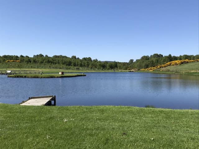 A stocked fishing lake at Achagour with a wooden angling platform, surrounded by grass banks and woodland