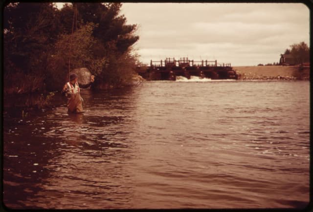 A wide stretch of Grand Lake Stream with an angler fly fishing near the bank and a dam spillway in the distance