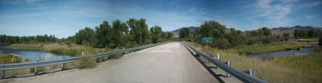 Wide view of a gravel road crossing the Clark Fork River on a bridge, with a road sign reading Clark Fork River, cottonwoods and mountains in the background