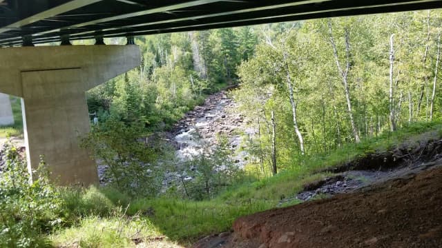 A rocky, boulder-strewn stretch of the Baptism River flowing through a wooded gorge near High Falls in Tettegouche State Park, Minnesota, seen from beneath a highway bridge.