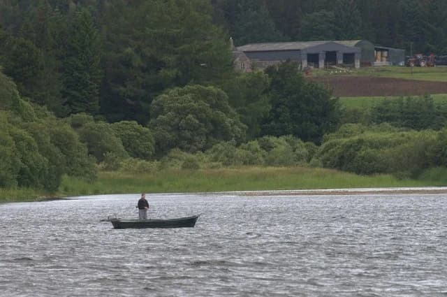 Wide view across Loch of Lintrathen reservoir in Angus, Scotland, with a fisherman at the water's edge and wooded hills beyond.