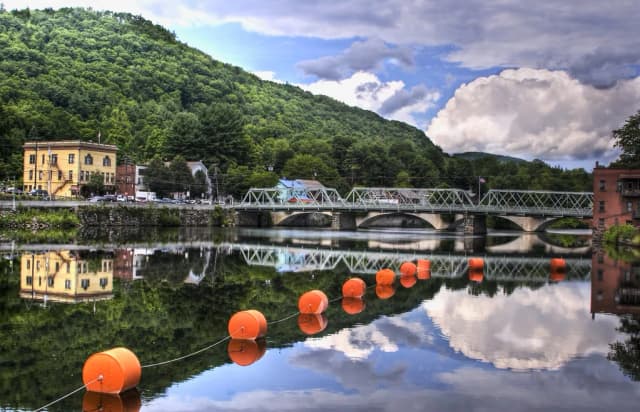 Still water on the Deerfield River reflecting a bridge, riverside buildings, and wooded hills in Massachusetts.