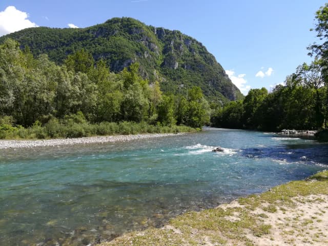 A wide clear river with turquoise water flowing over gravel past a forested limestone ridge in the Pyrenees
