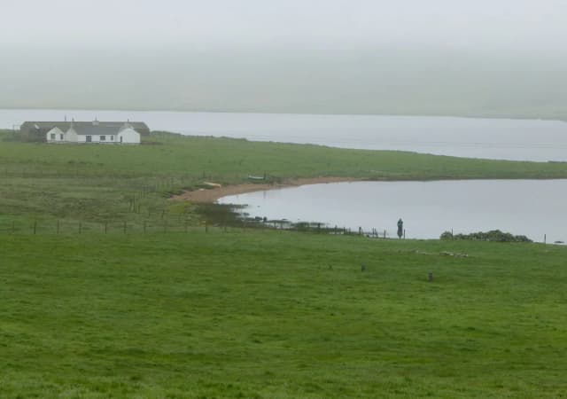A lone fisherman casts from the shore of the Loch of Spiggie in Shetland, with open water and low hills in the background.