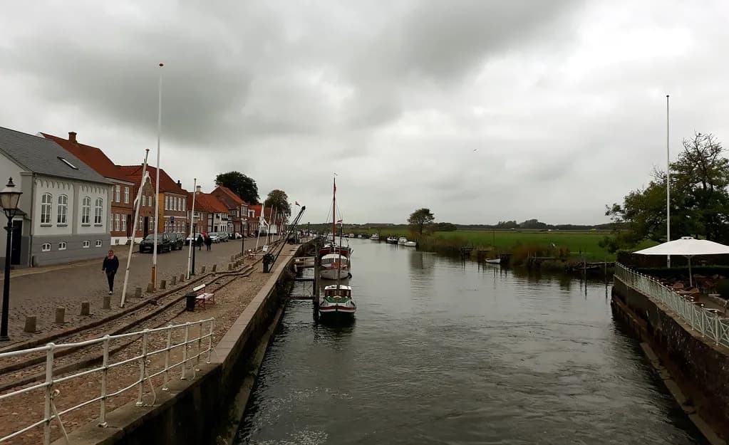 The Ribe river running past moored boats and traditional red-roofed houses in the old town of Ribe, Denmark, with green meadows in the distance.