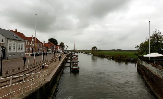 The Ribe river running past moored boats and traditional red-roofed houses in the old town of Ribe, Denmark, with green meadows in the distance.