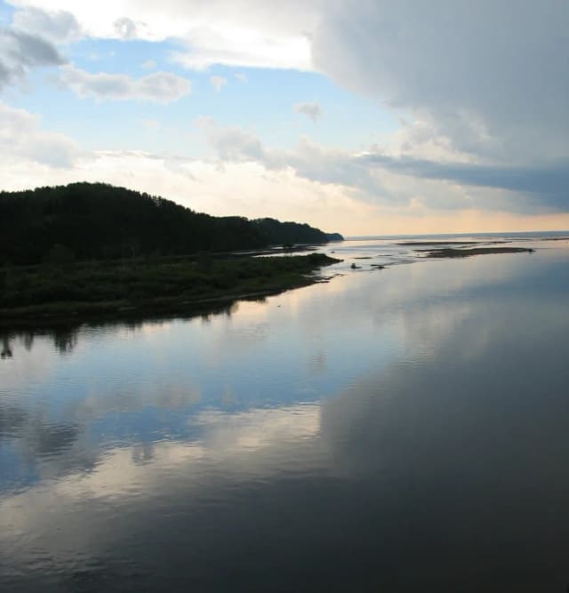 Wide calm river mouth reflecting a cloudy evening sky, forested shoreline on the left, Petite Cascapédia, Quebec.