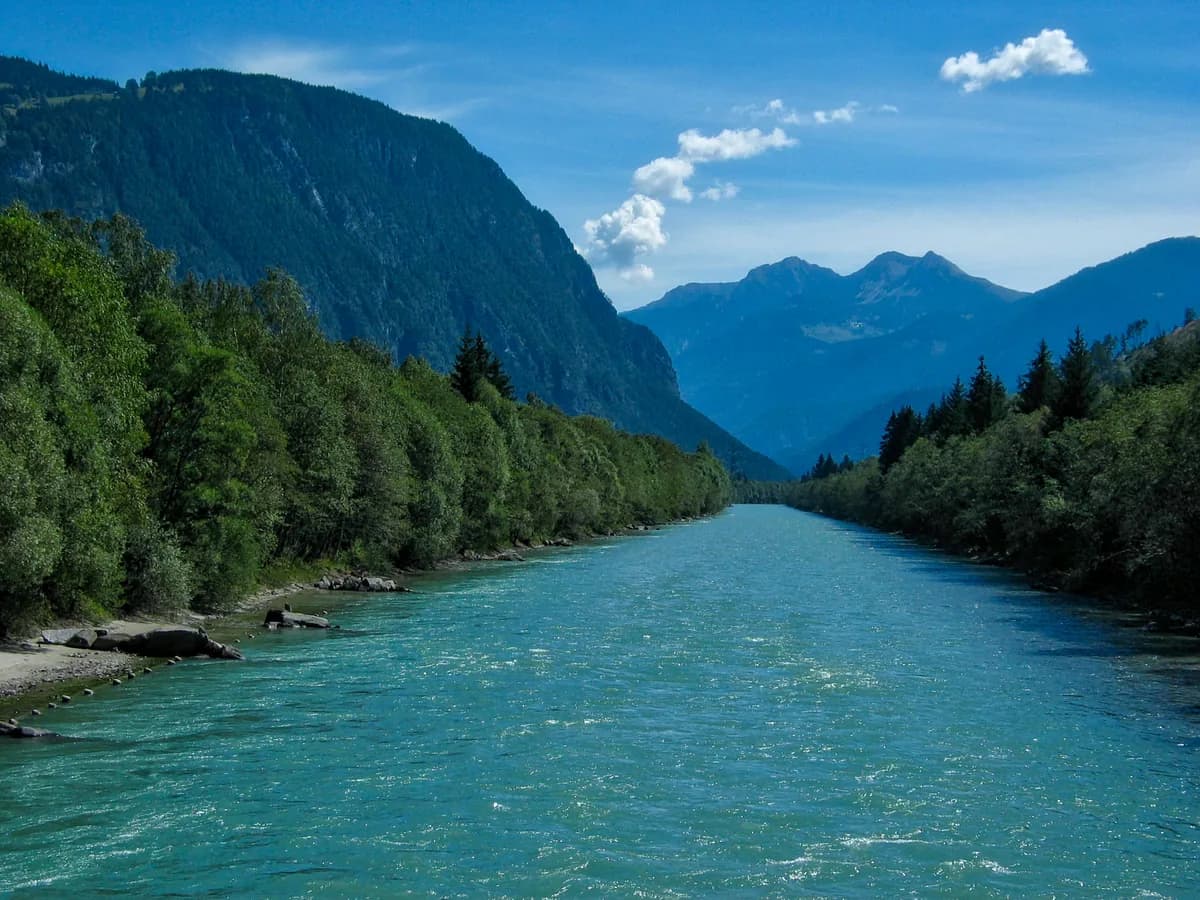 The turquoise Drau river flowing through a wide Alpine valley near Lienz, East Tyrol, with forested slopes and mountain peaks behind.