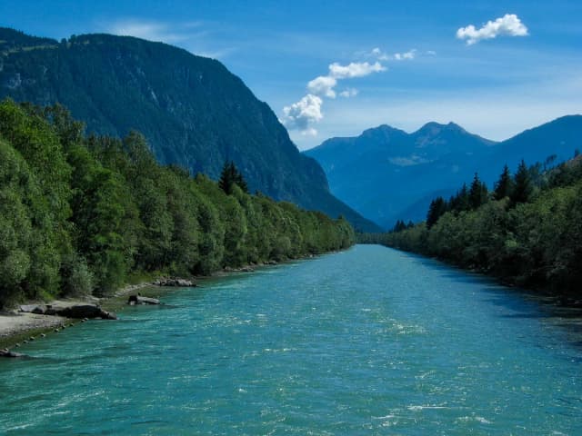 The turquoise Drau river flowing through a wide Alpine valley near Lienz, East Tyrol, with forested slopes and mountain peaks behind.