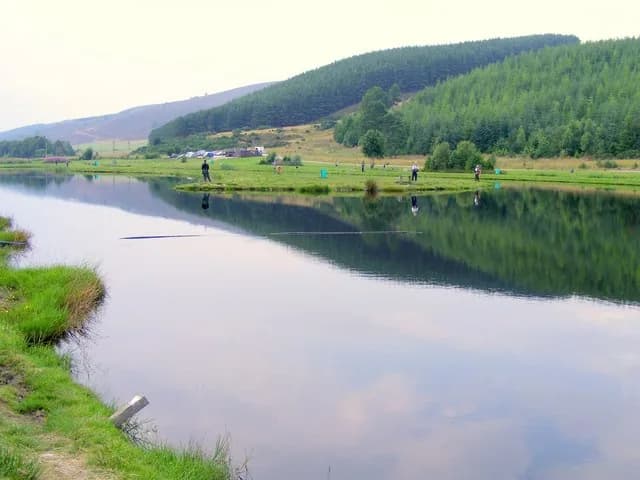 Wide view of a calm fishing pool at Glen of Rothes trout fishery, with anglers on the far bank and wooded hills beyond