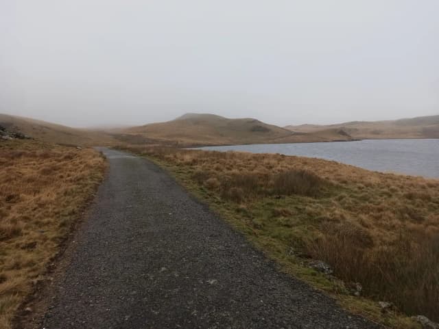 A gravel track beside Llyn Egnant, an upland lake in the Cambrian Mountains, Wales, with misty hills in the background.