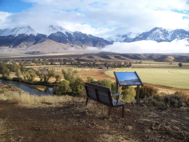 A river meanders through a wide agricultural valley with a viewing bench and interpretive sign in the foreground and snow-covered mountains behind