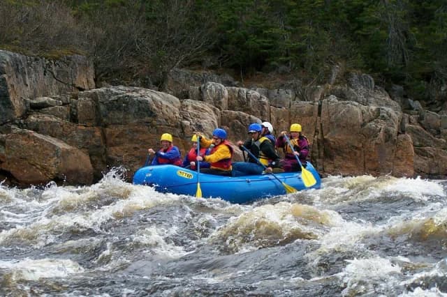 A blue raft carrying paddlers through churning whitewater rapids in a rocky river gorge, Main River, Newfoundland.