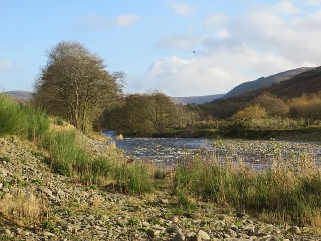 A wide, shallow stretch of Yarrow Water flowing over gravel with a stony bank in the foreground and hills rising beyond.
