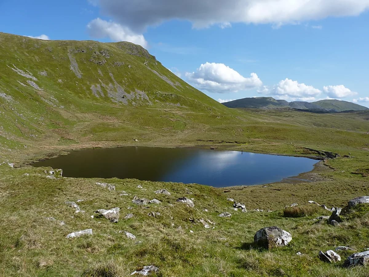 A small dark upland lake reflecting the sky, surrounded by grassy hillside and scattered rocks, with a hill rising behind it.
