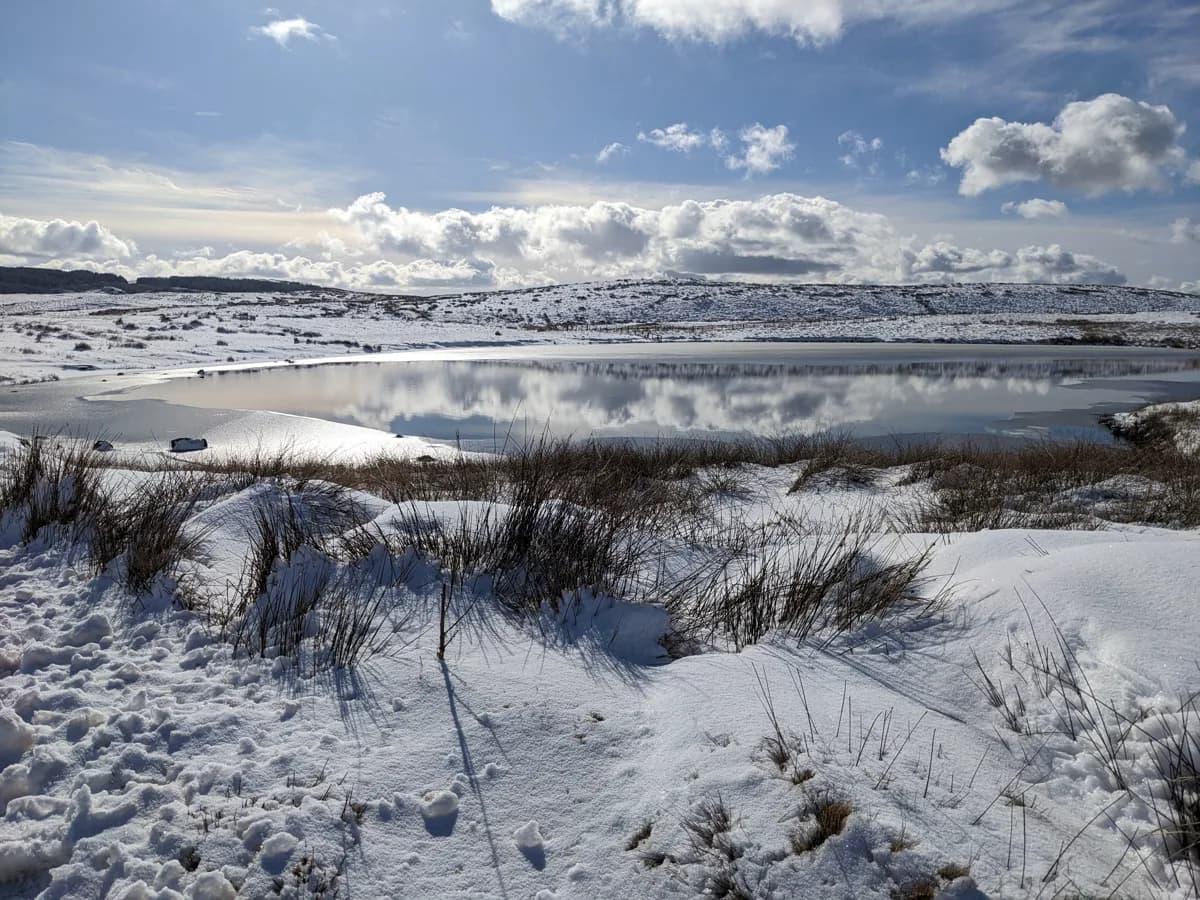 Snow-covered upland lake (Llyn Dubach y Bont) in Snowdonia with open water reflecting a partly cloudy sky.