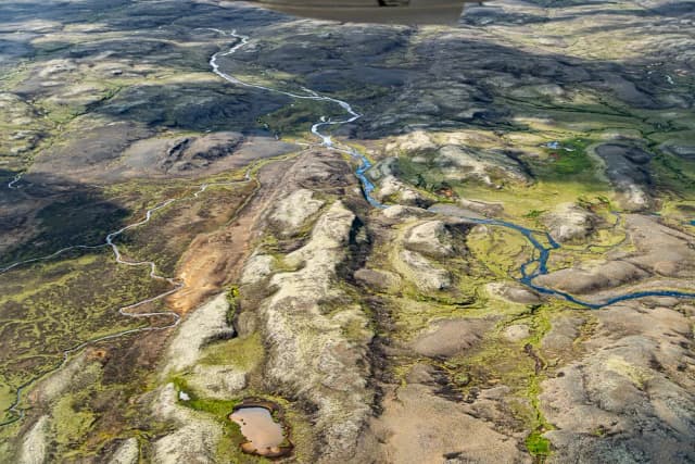 Aerial view of the Stora-Laxa river winding as a blue ribbon through green and grey volcanic lava-field terrain in Iceland.