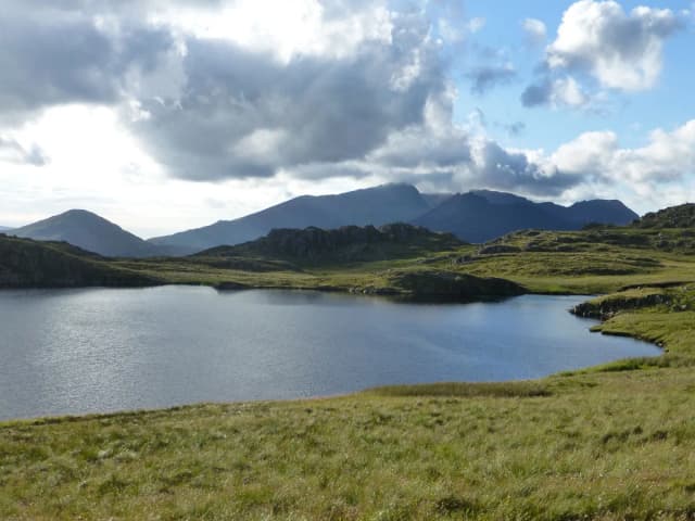 A calm upland lake surrounded by grassy moorland with rocky outcrops, with the mountains of Snowdonia including Snowdon visible under a partly cloudy sky.