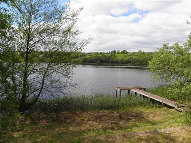 View across the calm water of Corranny Lough towards a wooden fishing jetty and wooded far shore
