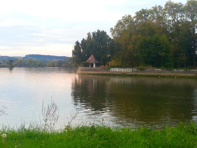 A wide, calm river at the confluence of the Adour and the Gaves Réunis, with trees and a small riverside gazebo marked Bec du Gave in the distance under an evening sky.