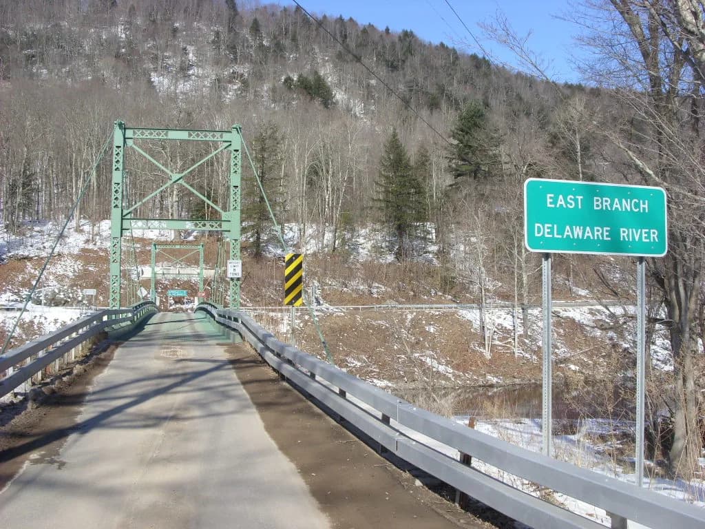Road sign reading East Branch Delaware River beside a green steel suspension bridge, with river water visible below, Catskills, New York.