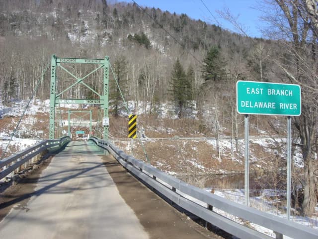 Road sign reading East Branch Delaware River beside a green steel suspension bridge, with river water visible below, Catskills, New York.