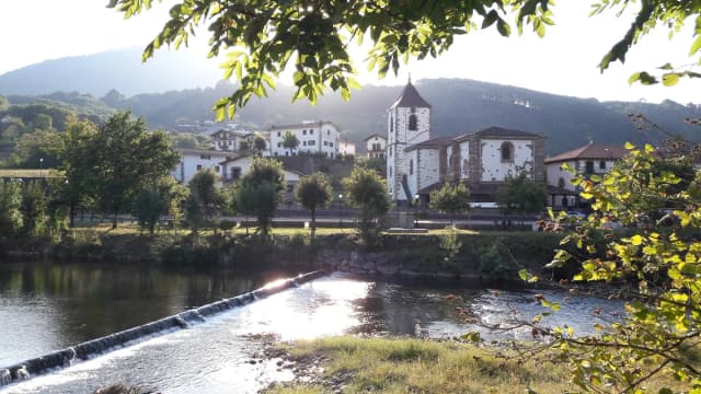 A river weir in the foreground with a Basque village of white houses and a church tower on the far bank, backed by wooded hills