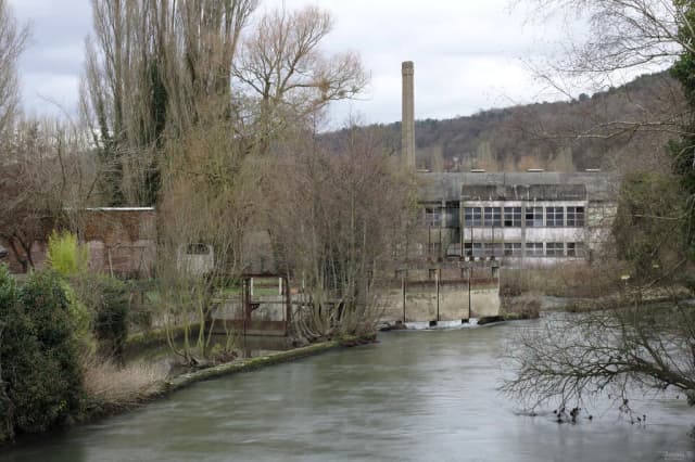 A clear, gently flowing river passing an old stone mill building in a wooded Normandy valley.