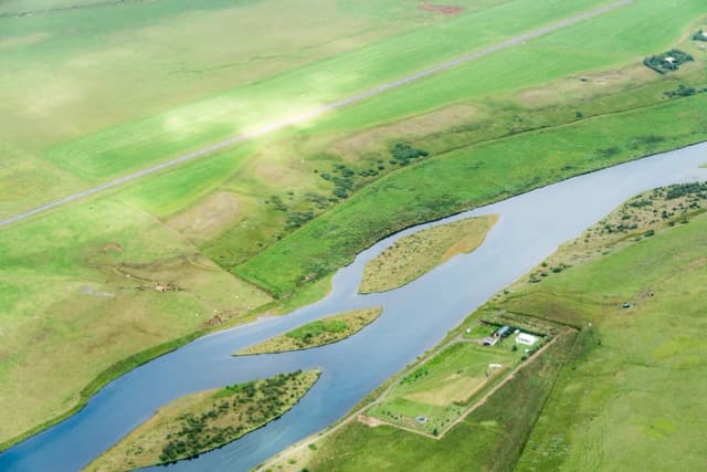 Aerial view of a wide, meandering glacial river with small vegetated islands, flowing through green Icelandic farmland near Hella.