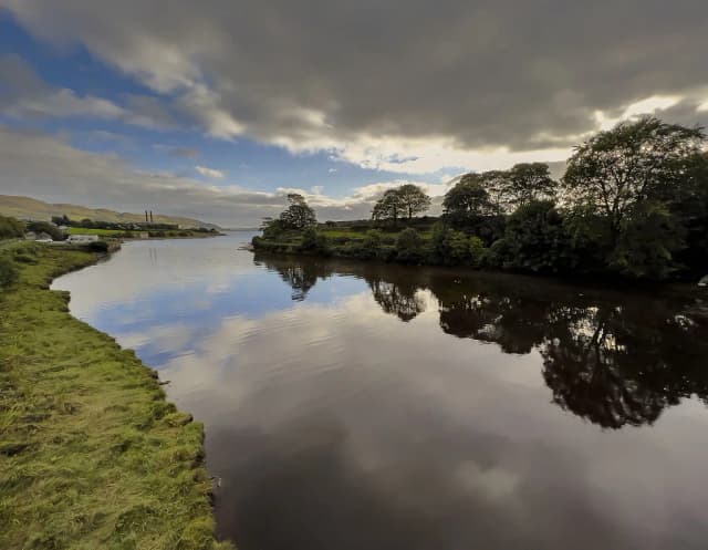 Still, dark water of the Crana River reflecting trees and sky near Buncrana, Co. Donegal, Ireland.