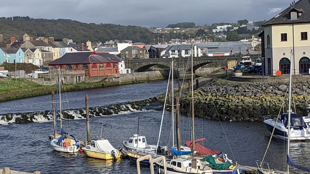River weir and stone bridge on the Afon Rheidol at Aberystwyth harbour, Wales, with moored boats and terraced houses beyond.