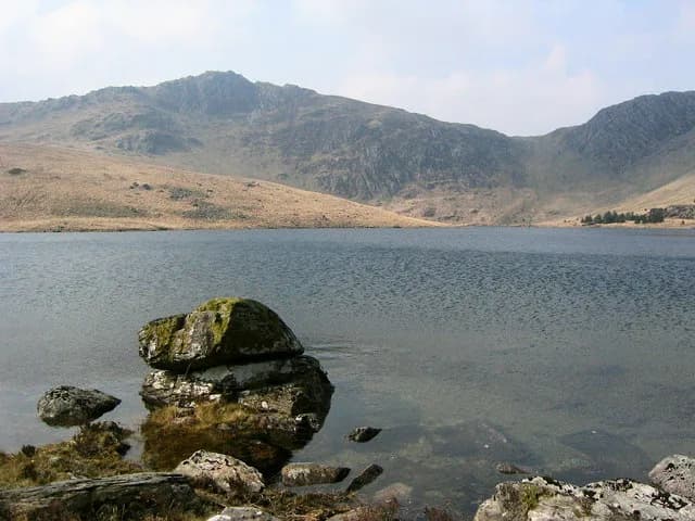 A moss-covered rock in the foreground of a calm upland lake, with bare mountain slopes rising behind under a clear sky.