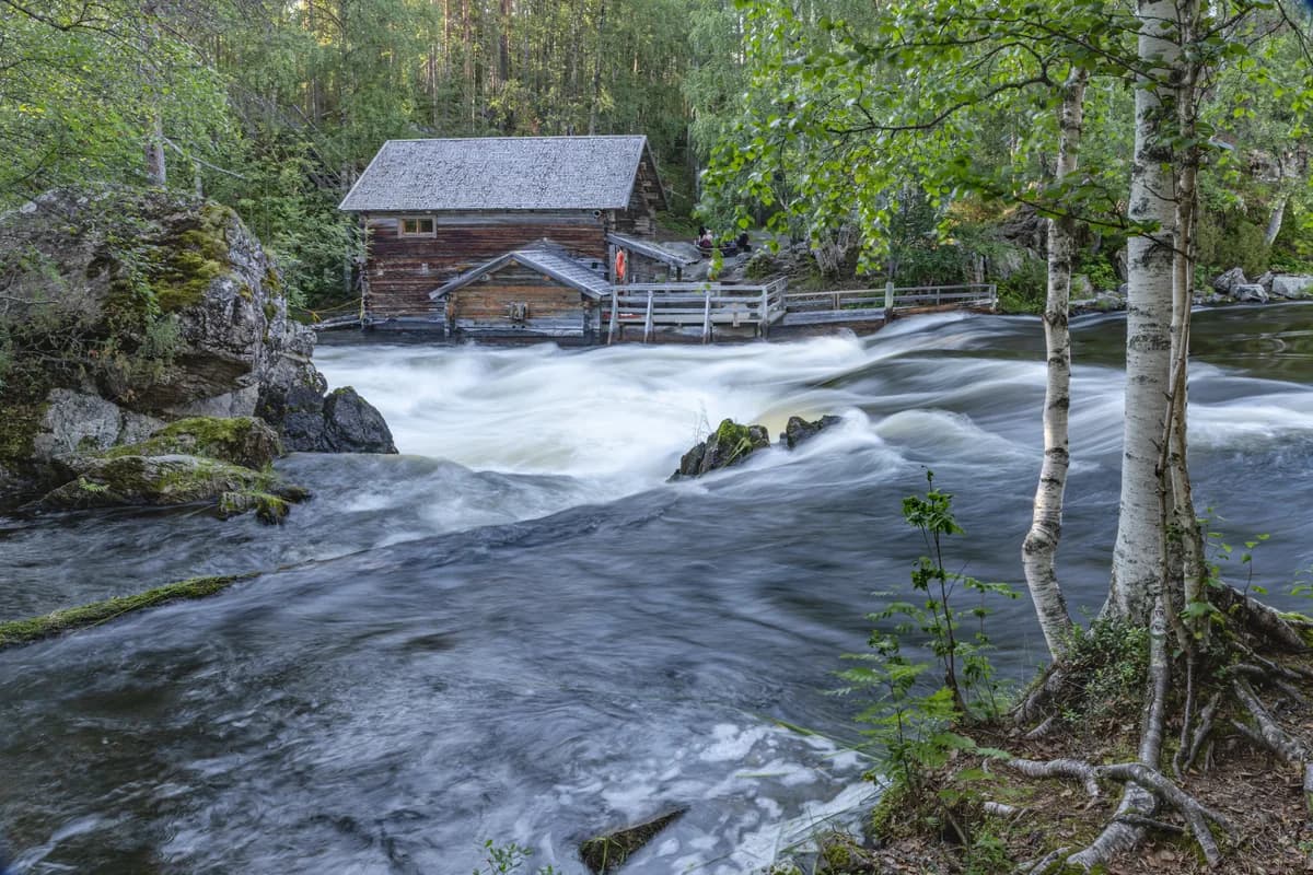 Wide view of whitewater rapids on the Kitkajoki River rushing past a historic wooden mill building in Kuusamo, Finland