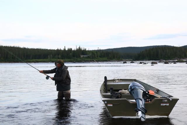An angler wades the wide Eagle River in Labrador, casting a fly rod beside a jon boat, with boreal forest along the far bank.