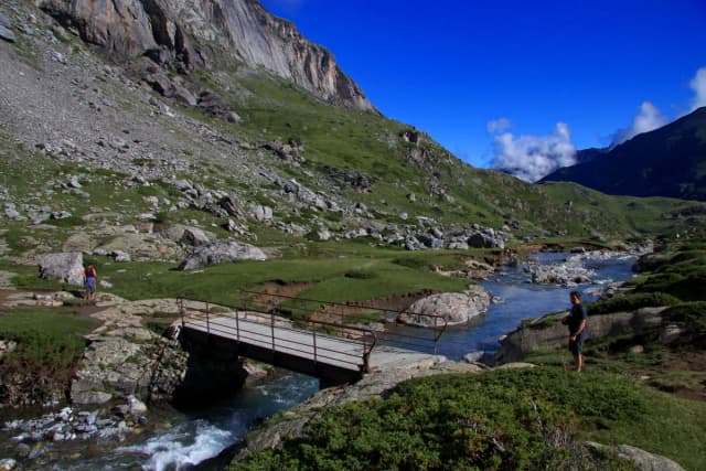 A mountain stream flowing through a rocky Pyrenean valley below a limestone cliff, crossed by a small footbridge