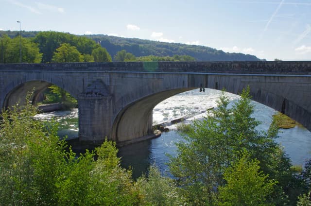 Stone arched bridge crossing the River Ain over a weir at Neuville-sur-Ain, with wooded hills behind.